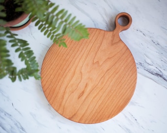 May include: A round wooden cutting board with a handle. The board is made of light brown wood and has a natural grain pattern. The handle is a simple loop.