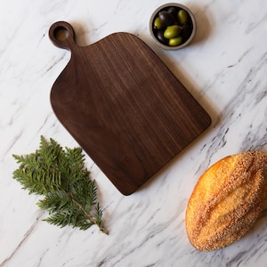 K&ouml;nnte beinhalten: Ein dunkelbraunes Holzschneidebrett mit Griff, neben einem Zweig gr&uuml;nen Laubes, einem Brotlaib und einer kleinen Schale Oliven. Die Artikel sind auf einer wei&szlig;en Marmoroberfl&auml;che angeordnet.