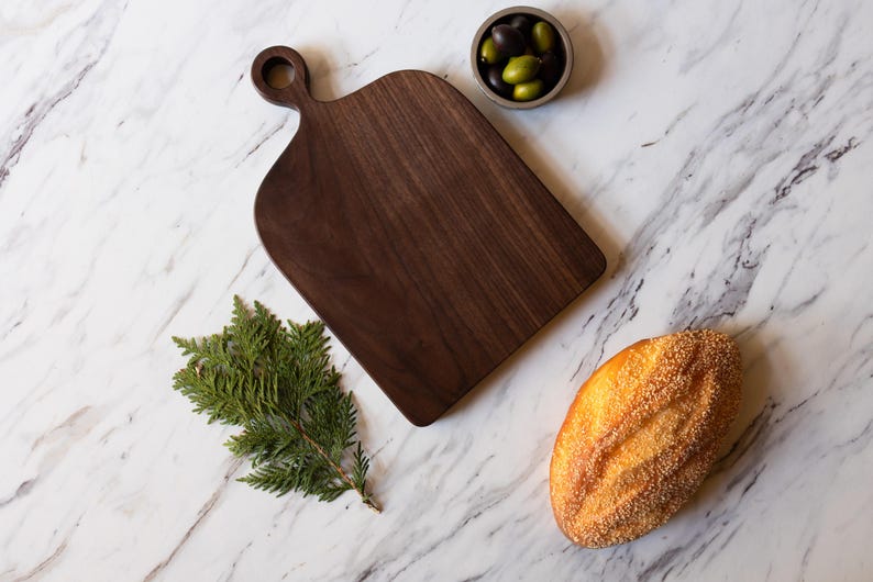 K&ouml;nnte beinhalten: Ein dunkelbraunes Holzschneidebrett mit Griff, neben einem Zweig gr&uuml;nen Laubes, einem Brotlaib und einer kleinen Schale Oliven. Die Artikel sind auf einer wei&szlig;en Marmoroberfl&auml;che angeordnet.