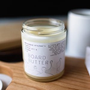 May include: A clear glass jar with a white label that reads "Mirondack Kitchen's Organic Coconut Oil & Beeswax Board Butter". The jar is filled with a white, creamy substance.