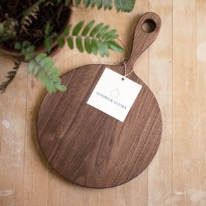 Pode incluir: T&aacute;bua de cortar redonda de madeira de noz com uma al&ccedil;a. A t&aacute;bua tem um acabamento de madeira natural e &eacute; etiquetada como "Adirondack Kitchen".