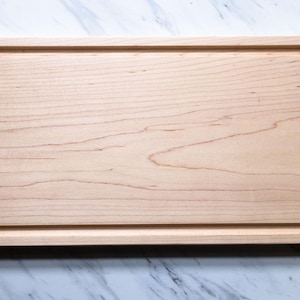 May include: A rectangular light-colored wooden cutting board featuring a smooth, flat surface and a recessed juice groove running along the entire inner perimeter. The wood grain is visible across the board, showcasing natural light brown tones.