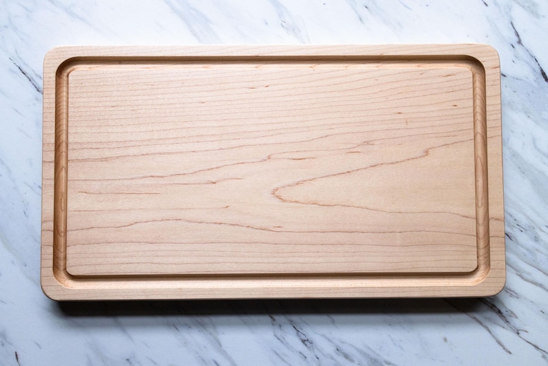 May include: A rectangular light-colored wooden cutting board featuring a smooth, flat surface and a recessed juice groove running along the entire inner perimeter. The wood grain is visible across the board, showcasing natural light brown tones.