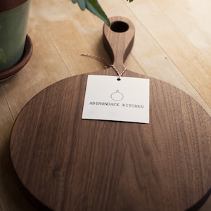Pode incluir: Uma t&aacute;bua de cortar redonda de madeira com uma al&ccedil;a. A t&aacute;bua &eacute; feita de madeira de noz e tem um acabamento natural. A t&aacute;bua &eacute; etiquetada com "Adirondack Kitchen".