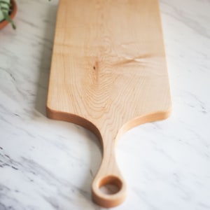 Puede incluir: Una tabla de cortar de madera natural con forma rectangular alargada y un solo mango con una abertura circular. La tabla es de madera de arce y tiene un color marr&oacute;n claro.
