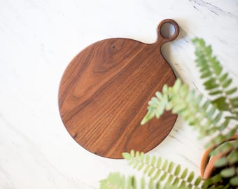 May include: A round wooden cutting board with a handle. The board is made of walnut wood and has a natural finish. The handle has a hole in it, which can be used to hang the board.