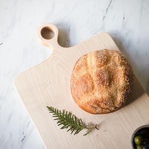 May include: A light wood cutting board with a unique handle, featuring a round loaf of bread with a golden crust and a sprig of green rosemary. A small bowl of olives is also on the board.