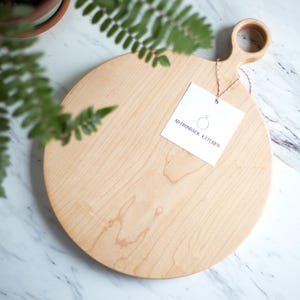 May include: A round wooden cutting board with a handle. The board is made of light wood and has a natural grain pattern. The handle is a simple loop and is attached to the board with a small piece of twine. The board is sitting on a white marble surface. A white tag with the text "Middleneck Kitchen" is attached to the handle.