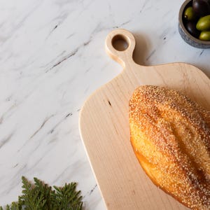 K&ouml;nnte beinhalten: Ein Brotlaib mit Sesamkruste liegt auf einem hellen Holzschneidebrett. Eine kleine Sch&uuml;ssel mit Oliven befindet sich in der N&auml;he. Der Hintergrund ist eine wei&szlig;e Marmoroberfl&auml;che mit dunkelgrauer Maserung.