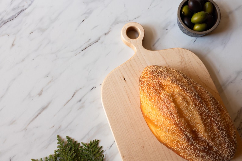 K&ouml;nnte beinhalten: Ein Brotlaib mit Sesamkruste liegt auf einem hellen Holzschneidebrett. Eine kleine Sch&uuml;ssel mit Oliven befindet sich in der N&auml;he. Der Hintergrund ist eine wei&szlig;e Marmoroberfl&auml;che mit dunkelgrauer Maserung.