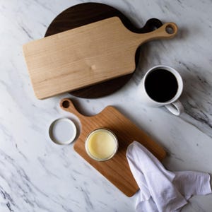 May include: Two wooden cutting boards, one with a dark wood finish and one with a light wood finish, are on a white marble surface. A small white cup with coffee is on the surface. A small jar of yellow butter is on the light wood cutting board.