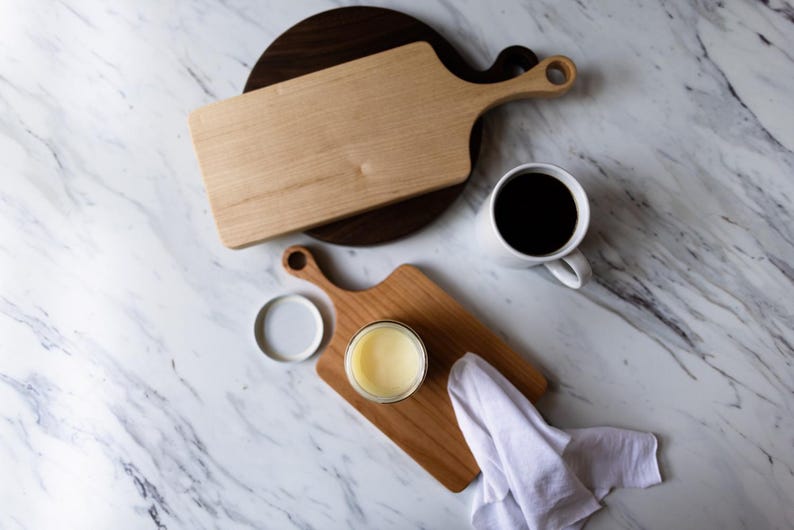 May include: Two wooden cutting boards, one with a dark wood finish and one with a light wood finish, are on a white marble surface. A small white cup with coffee is on the surface. A small jar of yellow butter is on the light wood cutting board.