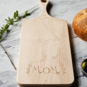 May include: A light wood cutting board with a handle and the word "MOM" engraved on the surface. The board is surrounded by a white marble surface.