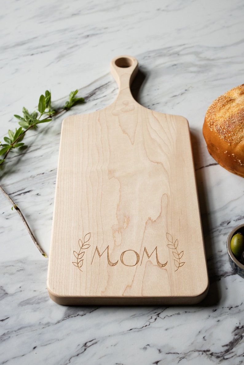 May include: A light wood cutting board with a handle and the word "MOM" engraved on the surface. The board is surrounded by a white marble surface.