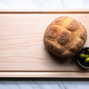 May include: A rectangular light-colored wooden cutting board with a grooved perimeter edge. A round, golden-brown crusty loaf of bread sits on the board next to a small dark bowl filled with a mix of green and black olives.