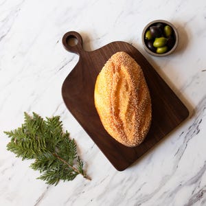 K&ouml;nnte beinhalten: Ein Brotlaib mit goldener Kruste und Samen liegt auf einem dunklen Holzschneidebrett. Eine kleine Schale mit Oliven und ein Zweig Gr&uuml;nzeug sind ebenfalls vorhanden, alles vor einem Marmorhintergrund.