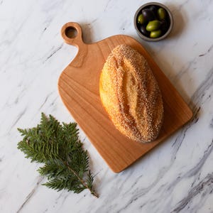K&ouml;nnte beinhalten: Ein Holzschneidebrett mit Griff h&auml;lt einen Laib Brot, der mit Sesamk&ouml;rnern bedeckt ist. Eine kleine Sch&uuml;ssel mit Oliven und ein Zweig Gr&uuml;nzeug liegen in der N&auml;he auf einer Marmoroberfl&auml;che. Das Brot ist goldbraun.