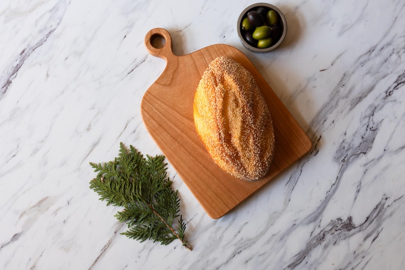 K&ouml;nnte beinhalten: Ein Holzschneidebrett mit Griff h&auml;lt einen Laib Brot, der mit Sesamk&ouml;rnern bedeckt ist. Eine kleine Sch&uuml;ssel mit Oliven und ein Zweig Gr&uuml;nzeug liegen in der N&auml;he auf einer Marmoroberfl&auml;che. Das Brot ist goldbraun.