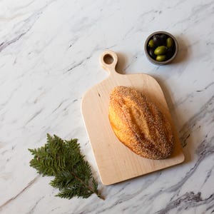K&ouml;nnte beinhalten: Ein h&ouml;lzernes Schneidebrett mit einem Brotlaib, einer kleinen Sch&uuml;ssel mit Oliven und einem Zweig Gr&uuml;n auf einer Marmoroberfl&auml;che. Das Brot ist goldbraun mit einer strukturierten Kruste. Die Oliven sind gr&uuml;n und schwarz.