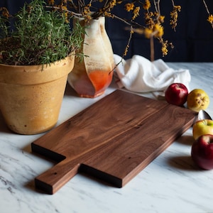 May include: A dark walnut wood cutting board with a handle. The board is rectangular with a handle on one end. A terracotta pot with a green plant, a vase, apples, and a fork are also in the image.