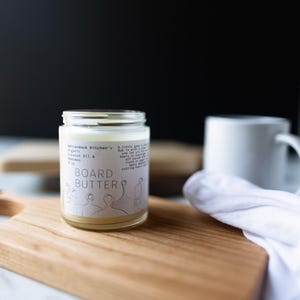 May include: A clear glass jar with a white label that reads "Adirondack Kitchen's Organic Coconut Oil & Beeswax BOARD BUTTER". The jar is sitting on a wooden cutting board.