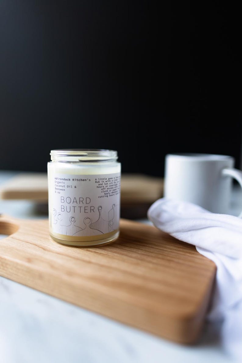 May include: A clear glass jar with a white label that reads "Adirondack Kitchen's Organic Coconut Oil & Beeswax BOARD BUTTER". The jar is sitting on a wooden cutting board.