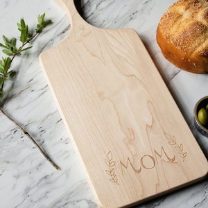 May include: A natural wood cutting board with a handle, engraved with the word "MOM" and leafy sprigs. The board is on a white marble surface.