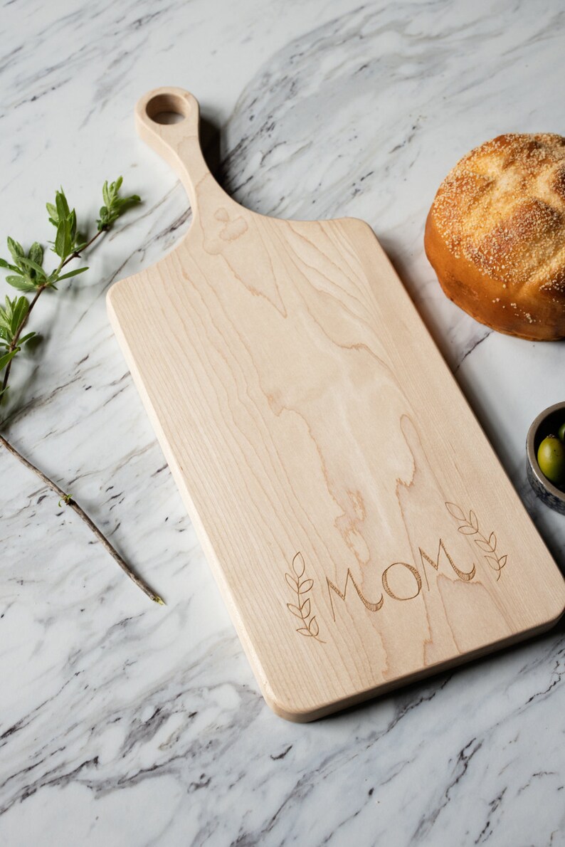 May include: A natural wood cutting board with a handle, engraved with the word "MOM" and leafy sprigs. The board is on a white marble surface.