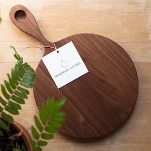 Pode incluir: Uma t&aacute;bua de cortar redonda de madeira com uma al&ccedil;a. A t&aacute;bua &eacute; de madeira escura e tem um acabamento natural. A t&aacute;bua tem um di&acirc;metro de 30 cm. Uma etiqueta branca com o texto "Adirondack Kitchen" est&aacute; presa &agrave; al&ccedil;a.