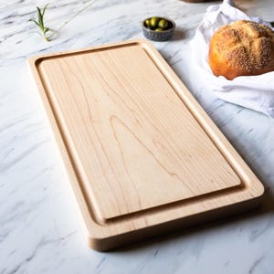 May include: A rectangular wooden cutting board with a juice groove. The board is made of light-colored wood and has a smooth surface. The cutting board is on a white marble surface. A small bowl of olives and a bread roll are in the background.