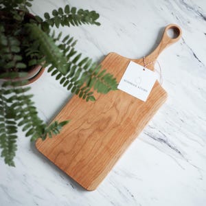 May include: A rectangular wooden cutting board with a handle. The board is made of light wood with a natural grain pattern. The handle is a simple loop shape. The board has a white tag attached to it with the text "Ashmore Kitchen".