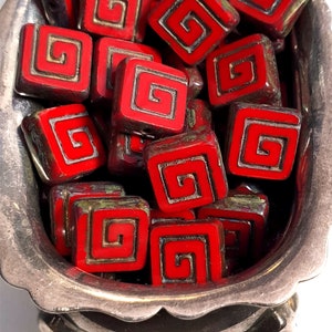 May include: A bowl filled with red and gold square-shaped beads with a swirling design. The beads are in a silver bowl with three feet.