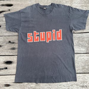 May include: A charcoal grey short-sleeve t-shirt featuring the word "stupid" printed in bold, block-style orange letters across the centre of the chest. The garment has a classic crew neckline and a straight hemline, showcasing a simple design.
