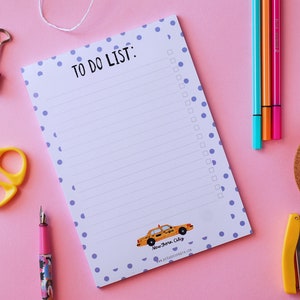 Può includere: Un blocco note bianco con pois viola e un'intestazione di lista di cose da fare. Il blocco note presenta un'illustrazione di un taxi giallo con il testo "New York City" in basso. Il blocco note è su una superficie rosa con vari articoli da ufficio, tra cui forbici, una graffatrice e penne.