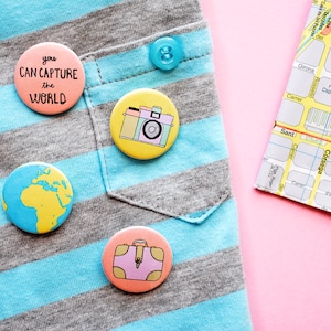 May include: A blue and grey striped shirt with four colourful pinback buttons. The buttons feature a globe, a camera, a suitcase, and text that reads "you can capture the world".