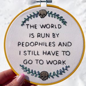 May include: Embroidered hoop art with the text "THE WORLD IS RUN BY PEDOPHILES AND I STILL HAVE TO GO TO WORK." The design includes a skull and leaf accents. The hoop is yellow.