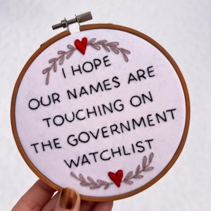 May include: Embroidered hoop art with the text "I HOPE OUR NAMES ARE TOUCHING ON THE GOVERNMENT WATCHLIST". The design features a white background, black lettering, a wooden hoop, and red heart accents.