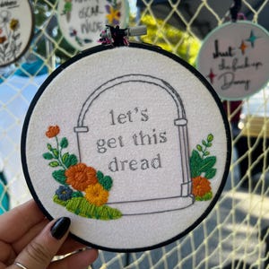 May include: Embroidered hoop art with the phrase "let's get this dread" in gray lettering. The design includes a tombstone shape with orange and yellow flowers and green foliage. Other hoops with text and floral designs are visible.