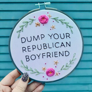 May include: Embroidered hoop art with the text "DUMP YOUR REPUBLICAN BOYFRIEND" in black thread. The design includes pink and gold floral accents and green leaves, all on a white background. The hoop is held by a hand.