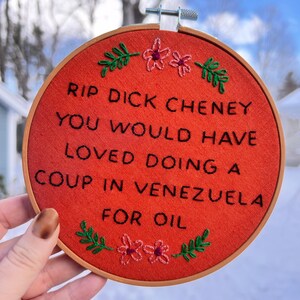 May include: A round, orange fabric embroidery hoop with black text that reads "RIP DICK CHENEY YOU WOULD HAVE LOVED DOING A COUP IN VENEZUELA FOR OIL." The design includes embroidered green leaves and pink flowers.