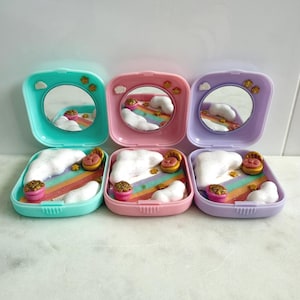 May include: Three open compact mirrors in pastel shades: turquoise, pink, and lilac. Each mirror contains a miniature scene with a rainbow, clouds, and small decorative features. The compacts are square with rounded corners and a small mirror.