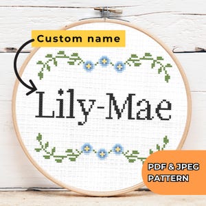 May include: A cross-stitch embroidery hoop with the name "Lily-Mae" in black thread. The design includes a floral border with green leaves and blue flowers. The text "Custom name" is above the name, and "PDF & JPEG PATTERN" is in the corner.