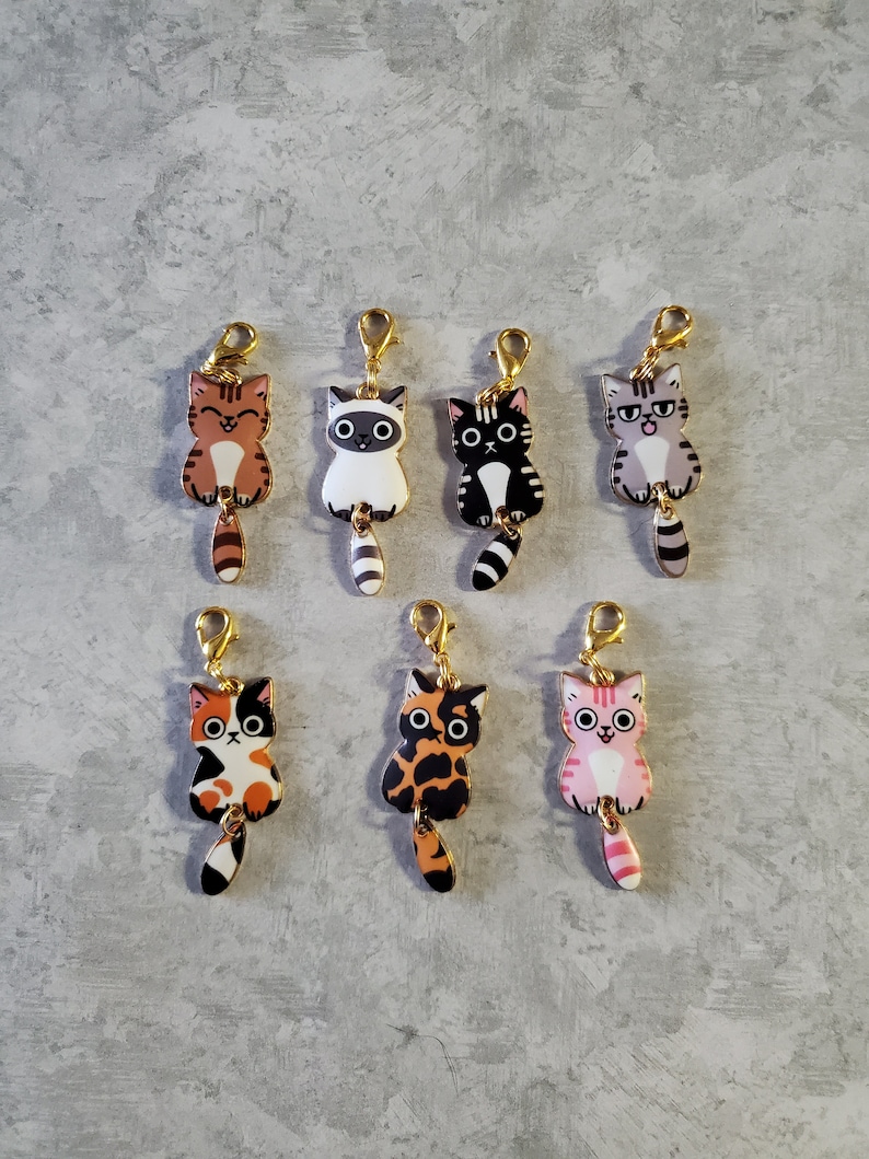 May include: Six colourful cat charms with gold swivel clips. Each charm features a different cat design with a unique tail.