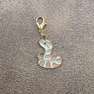 May include: A gold-coloured metal charm in the shape of a pink and white snake with a gold clasp.