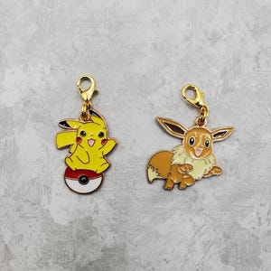 May include: Two gold-colored metal charms featuring Pokemon characters. The first charm features Pikachu standing on a Pokeball. The second charm features Eevee.