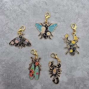 May include: Five insect-themed charms with gold-coloured clasps. The charms include a moth, a bee, a beetle, a pair of shoes, and a scorpion, all with black enamel and floral accents in various colours.
