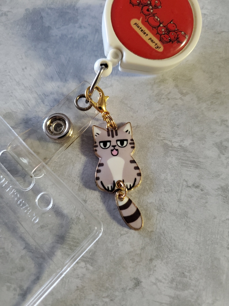 May include: A gold retractable badge holder with a red and white circular charm that says "platelet party!" and a grey and black cat charm with a gold chain.