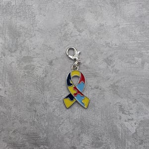 Puede incluir: Un charm de cinta colorida con un cierre plateado. La cinta está diseñada con patrones de piezas de rompecabezas en rojo, amarillo, azul y negro. El charm es probablemente un símbolo de concienciación sobre el autismo.