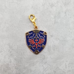 May include: A gold-colored metal charm featuring the iconic Triforce symbol from the Legend of Zelda video game series. The charm is shaped like a shield with a blue background and red Triforce design. It has a gold-colored clasp for attaching to a bag or keychain.