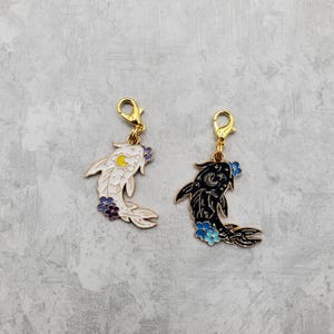 May include: Two gold-tone metal charms shaped like koi fish. One fish is white with yellow and blue accents, the other is black with blue accents. Both charms have a gold-tone clasp.